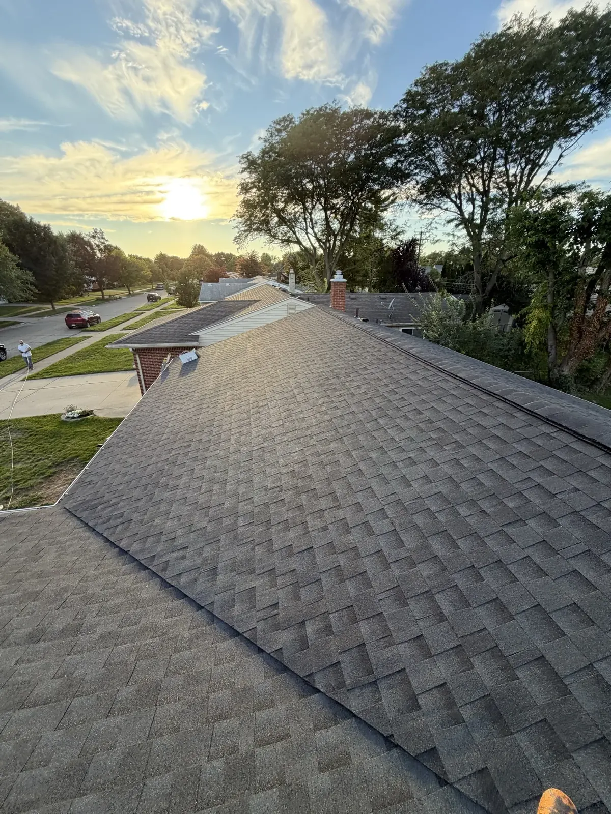 A finished gray shingle roof at sunset above a tree-lined residential street